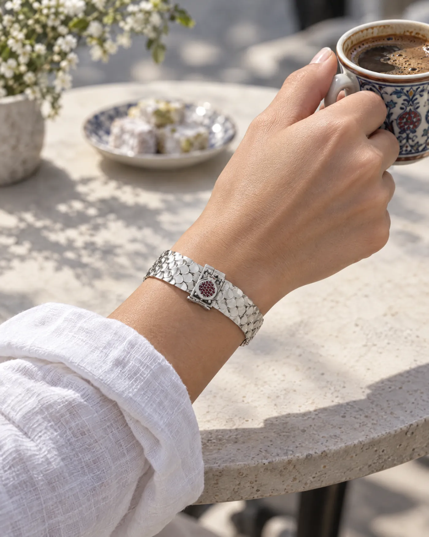 Slim fish-scale silver bracelet with a garnet pomegranate clasp on a wrist holding an Armenian coffee cup