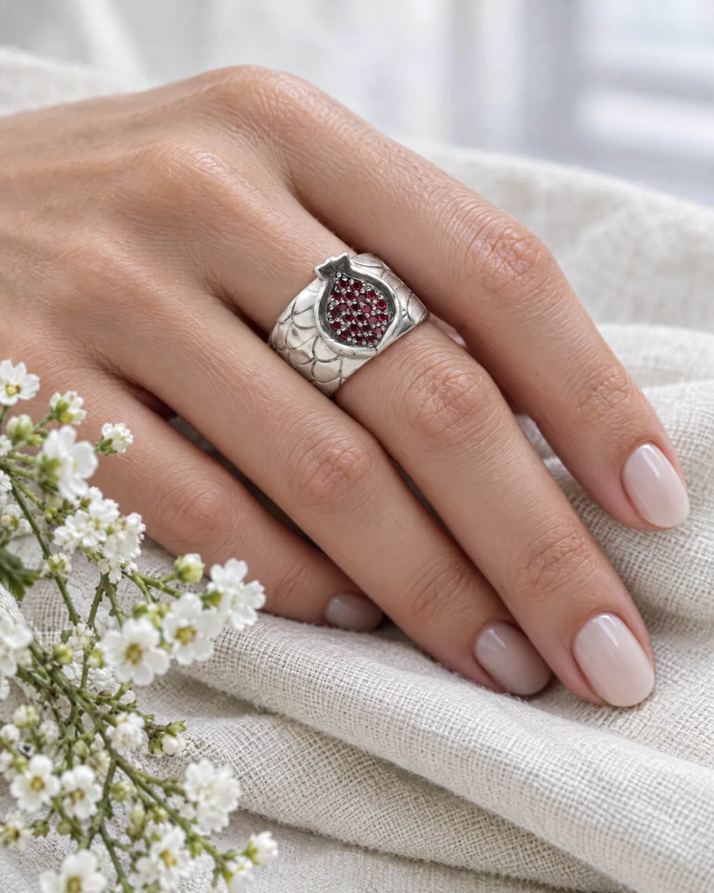 Silver ring with a garnet pomegranate top and fish-scale band on a hand resting on linen